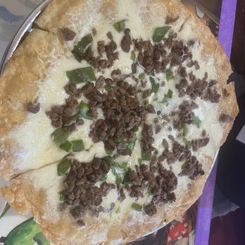 Taco Giro - A pizza featuring a golden, crispy crust with a layer of melted cheese, topped with cooked ground beef and chopped green peppers. The pizza is served on a metal tray, with some cheese and toppings visible on the edges. The background includes a colorful tablecloth and part of a person's arm, indicating a casual dining setting. - Breakfast & Brunch Near Me - Apache Junction, Arizona