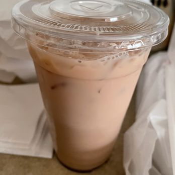 Cafe Fresco - A tall, clear plastic cup filled with iced coffee or tea, showing a light brown color with visible ice cubes inside. The cup has a transparent, sealed plastic lid with a circular design on top. The background includes a white napkin and some indistinct objects, suggesting a casual setting such as a cafe or fast-food restaurant. - Cafes Near Me - Crown Point, Indiana