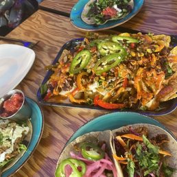 Dylan Taco - The image shows a wooden table set with multiple Mexican dishes, including tacos with toppings like jalapeños and pickled onions, a large plate of nachos with vegetables and sliced jalapeños, and a salad with greens and tomatoes. There are also empty plates, utensils, and a drink bottle visible, indicating a meal in progress at Proper Taco.