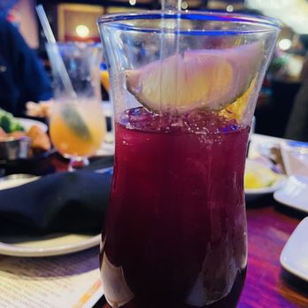 Jasper's - White Moscato Sangria and Sweet Red Sangria - A tall glass of red sangria with a lemon wedge garnish is on a restaurant table, with other drinks and dishes visible in the background. - American Near Me - Upper Marlboro, Maryland