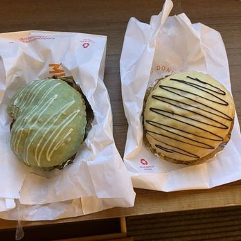 Rainbow Bakery - Matcha and mango cream puffs. So large and filled with flavored cream with a light icing. So delicious! - The image shows two cream puffs on a wooden table, each resting on a piece of white paper. The cream puff on the left has green icing with white drizzle, while the one on the right has a yellowish glaze with dark chocolate drizzle. Both pastries are displayed without additional packaging or utensils, highlighting their decorative toppings and filling. - Bakeries Near Me - Queens, New York