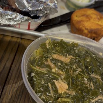 Test Kitchen BBQ - Collard greens - A clear plastic container holds cooked collard greens with visible stems and seasoning. In the background, there are foil-wrapped food items, fried plantains, and small lime wedges on a table, suggesting a meal setting. The image emphasizes the collard greens as part of a diverse food spread. - Barbeque Near Me - Waukegan, Illinois