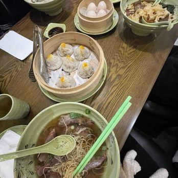 E Noodle - D1.Pork Soup Dumplings(6), shrimp balls - The image shows a table set with Chinese cuisine, including soup dumplings, noodle soup, and other dishes, with green ceramic bowls and chopsticks. - Noodles Near Me - Brooklyn, New York