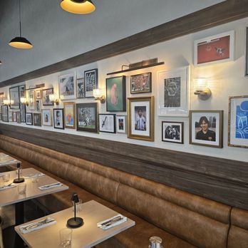 Keepers - The restaurant interior features a long brown leather banquette along a wall decorated with numerous framed photographs and artwork. Small wall-mounted lights illuminate the gallery wall. Several tables are set with utensils, glasses, and water bottles, ready for diners. Overhead, black pendant lights hang from the ceiling, providing additional lighting. The decor combines modern lighting with a cozy, artistic atmosphere. - American Near Me - Harrison, New Jersey