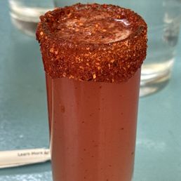 Mexican candy shot! - A tall, clear shot glass is filled with a red beverage, topped with a rim coated in chili powder or crushed red pepper. The drink appears to be a spicy cocktail or shot, with a vibrant red color and a textured, spicy rim. The background includes a glass of water and a blurred table surface, indicating a casual dining or bar setting.