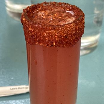 Centro Mexican Kitchen - Mexican candy shot! - A tall, clear shot glass is filled with a red beverage, topped with a rim coated in chili powder or crushed red pepper. The drink appears to be a spicy cocktail or shot, with a vibrant red color and a textured, spicy rim. The background includes a glass of water and a blurred table surface, indicating a casual dining or bar setting. - Mexican Near Me - Boulder, Colorado