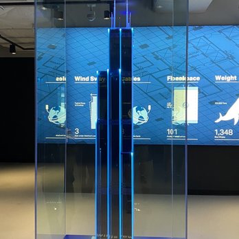 Skydeck Chicago - A tall, narrow display case with blue LED lighting runs vertically inside a glass enclosure. The display appears to be a technological or scientific exhibit, with a digital background featuring diagrams and data related to wind, cables, and weight. The setting suggests a modern, high-tech environment, possibly a museum or science center. - Observatories Near Me - Chicago, Illinois