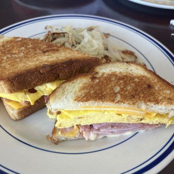 Waffle Pancake House - Moon Over Miami Breakfast sandwich. Ham, American Cheese, White American Cheese and scrambled eggs on sourdough bread. - A breakfast sandwich featuring ham, scrambled eggs, and American cheese between two slices of toasted bread is presented on a white plate with blue trim. The sandwich is cut in half, revealing the layers of ingredients. Behind the sandwich, there is shredded cabbage, and in the background, other plates with food are visible on a dark table. - Breakfast & Brunch Near Me - Chicago, Illinois