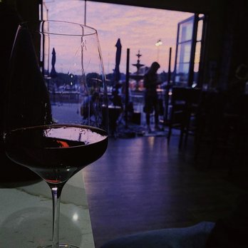 Apericena Wine Bar - Red wine - A glass of red wine is positioned on a table inside a restaurant, with a view of an outdoor patio through an open window. The patio features umbrellas, a fountain, and a person standing near the railing. The scene is set during sunset, with a sky displaying pink and purple hues. The interior appears dimly lit, emphasizing the silhouette of the outdoor scene. - Wine Bars Near Me - Upper Saint Clair, Pennsylvania