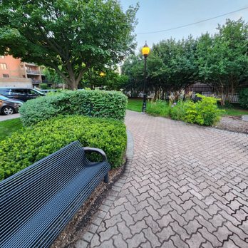 Moehling Park - The image shows a paved brick pathway in a park or garden area, lined with a black metal bench on the left side. Surrounding the path are well-maintained bushes, trees, and flowering plants. Two street lamps are visible along the path, providing lighting. In the background, parked cars and residential buildings can be seen, indicating an urban or suburban setting. The scene appears to be during early evening or late afternoon, with natural light and some street lighting. - Parks Near Me - Mount Prospect, Illinois