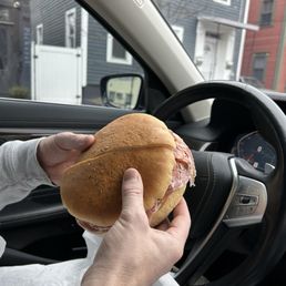 Muffuletta - A person is holding a large muffuletta sandwich with bread and meat inside a vehicle, with a view of residential buildings outside.