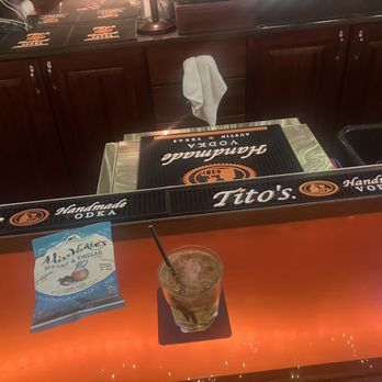 Rex Tavern - Jack And Coke - A cocktail with ice and a straw is positioned on a bar counter at Tito's bar, with a bag of Miss Vickie's sea salt & vinegar chips beside it. The bar features a dark wood finish, branded bar mats with Tito's Handmade Vodka, and a towel hanging on the back of a chair. The background shows additional bar surfaces and equipment, indicating a casual bar setting. - Sports Bars Near Me - Chicago, Illinois