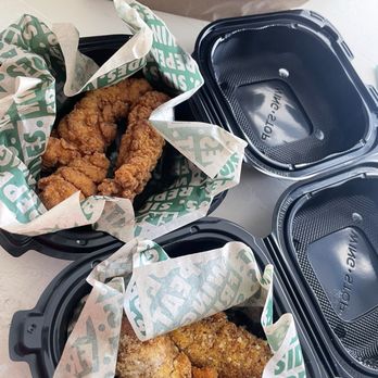 Wingstop - Two black plastic takeout containers filled with fried chicken pieces are lined with branded paper that has green and white text. The containers are open, showing the fried chicken inside. A paper bag with a business logo is visible in the background, placed on a white surface. The image suggests a meal from a fast-food or fried chicken restaurant. - Chicken Wings Near Me - Albany, New York
