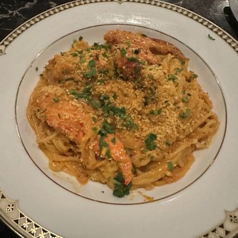 Geppetto - A plate of seafood pasta, likely lobster or shrimp, is presented on a decorative white plate with a gold and black border. The dish is garnished with chopped herbs and sprinkled with grated cheese. The background shows a dark table surface with utensils and glasses, indicating a restaurant environment. - Italian Near Me - Cambridge, Massachusetts