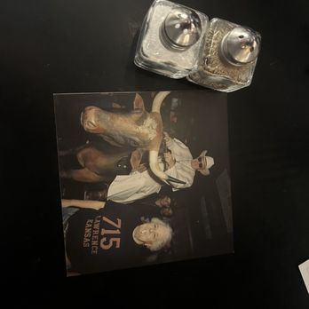 715 - Table - A photo shows a person with white hair wearing a black shirt with '715' and 'Kansas' printed on it, standing next to a dog, with salt and pepper shakers on a dark table. - Italian Near Me - Lawrence, Kansas