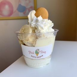 Banana Pudding - A banana pudding sundae in a clear plastic cup features whipped cream, banana slices, a cookie, and a logo sticker from Pink Pony Soft Serve.