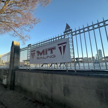 MIT Sailing Pavilion - A metal fence with a MIT Sailing banner is situated along a waterfront in Cambridge, MA, with boats and city buildings visible in the background. - Boating Near Me - Cambridge, Massachusetts