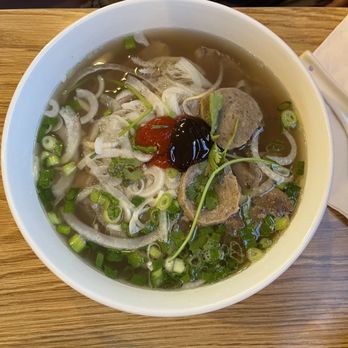 Việt kitchen - Pho Beef Noodle Soup - A large white bowl contains Vietnamese pho with clear broth, beef slices, chopped green onions, cilantro, and sliced onions. A dollop of red chili sauce and a dark sauce or hoisin are added on top. The bowl is placed on a wooden table, with a person's hand and part of their arm visible in the background. The setting appears to be a casual restaurant. - Vietnamese Near Me - Albuquerque, New Mexico