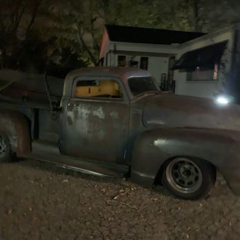 Terror Maze - A vintage pickup truck with a rusted, weathered exterior is parked outdoors on a dirt surface. The truck appears to be an older model, with visible rust and signs of age. It is positioned near a small house or building, with trees and other structures faintly visible in the background. The scene is dimly lit, with only a small amount of light illuminating parts of the truck and surroundings. - Haunted Houses Near Me - Springfield, Ohio