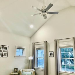 The image shows a bedroom with a high vaulted ceiling, a ceiling fan with a decorative chandelier, and three windows with curtains. The windows allow natural light into the room, and there is a small sitting area with a beige armchair, a side table, and a lamp. The room has a neutral color palette with white walls and light-colored furnishings.