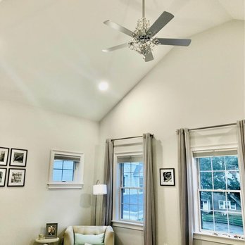 Kinney Electric - The image shows a bedroom with a high vaulted ceiling, a ceiling fan with a decorative chandelier, and three windows with curtains. The windows allow natural light into the room, and there is a small sitting area with a beige armchair, a side table, and a lamp. The room has a neutral color palette with white walls and light-colored furnishings. - Electricians Near Me