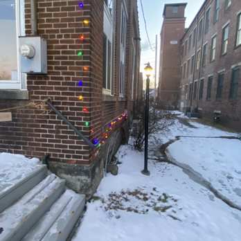 Drawing Board Brewing - a brick building with christmas lights on it - A narrow alleyway covered with snow runs between brick buildings. A vintage-style street lamp is positioned along the path, and colorful string lights are hung along the wall of the building on the left. The alley features a small set of stairs, a sign pointing to a can sale, and some bushes. The scene appears to be during late afternoon or early evening, with a partly cloudy sky. - Breweries Near Me - Florence, Massachusetts