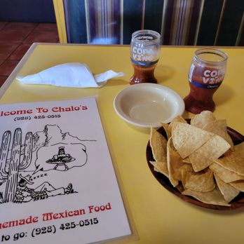 Chalo's - The image shows a yellow table with two glasses of beer, a bowl of tortilla chips, a napkin, and a menu from Chalo's Mexican restaurant. The menu features a cactus and mountain illustration with text welcoming customers and providing contact information. The background includes tiled flooring and a colorful wall or partition. - Mexican Near Me - Globe, Arizona