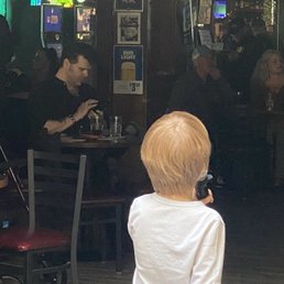 Thank you Embers for allowing children to participate in karaoke night!  He was adorable! - A child with blonde hair, wearing a white shirt, is holding a microphone in a dimly lit bar or restaurant setting.