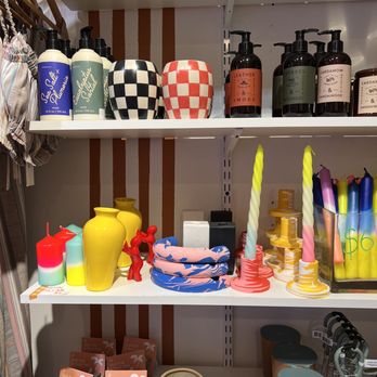 Poor George - Fun candles - Two white shelves contain various products including candles, vases, and personal care items. The top shelf has bottles of lotion and spray, along with checkered vases. The middle shelf displays colorful candles, vases, and decorative items, including twisted candles and small containers. The bottom shelf features boxed products, a candle holder, and other small accessories. The background shows a wall with vertical brown stripes, indicating a retail environment. - Women's Clothing Near Me - Cold Spring, New York
