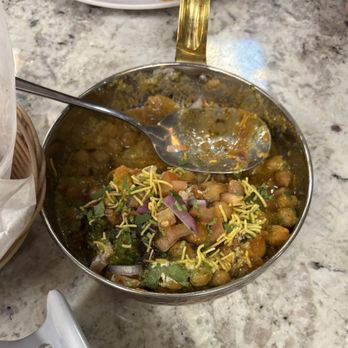 Tikka TapRoom - Chicken Biryani - A metal bowl filled with chicken biryani, featuring chickpeas and a yellowish sauce, is placed on a marble table. The dish is garnished with chopped red onions, cilantro, and crispy noodles on top. A spoon rests inside the bowl, and part of a basket with bread or naan is visible nearby. The setting appears to be a restaurant or dining area. - Indian Near Me - Lindenhurst, Illinois