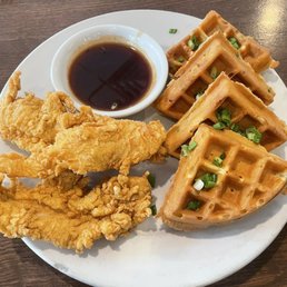 Chicken waffles - A plate features fried chicken, Belgian waffles topped with chopped green onions, and a small bowl of syrup, served on a wooden table.