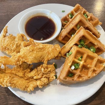 Pomegranate Restaurant - Chicken waffles - A plate features fried chicken, Belgian waffles topped with chopped green onions, and a small bowl of syrup, served on a wooden table. - American Near Me - Aurora, Illinois