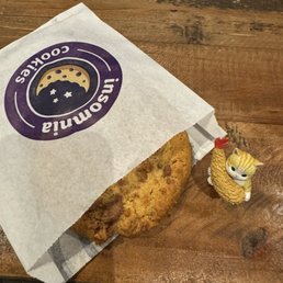 Chocolate Peanut Butter Cup - A fried chicken drumstick with a crispy, golden-brown coating is partially wrapped in a white paper bag featuring a purple logo that reads 'Insomnia Cookies' with a moon and stars graphic. The drumstick is placed on a wooden table with visible grain and texture. Next to the food item, there is a small figurine of a cat with a chicken design, including a red comb on its head, and a fluffy, textured body. The image is taken from a top-down perspective, showing the person's white shoes and part of their legs at the bottom of the frame.