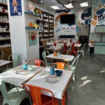 Color Me Mine - The image depicts the interior of Color Me Mine in Glenview, with several tables covered with white paper and art supplies, including paint palettes and brushes. Shelves along the walls display various painted pottery pieces, such as bowls and mugs. The walls are decorated with colorful, abstract murals and artwork, creating a lively atmosphere. The ceiling has a fan and multiple light panels, and a large TV is mounted on the wall. The space is organized for customers to paint and decorate ceramics. - Paint-Your-Own Pottery Near Me - Glenview, Illinois