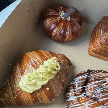 Baker 305 - The image features three pastries inside a cardboard box. The pastries include a croissant topped with white icing and crushed pistachios, a pumpkin-shaped pastry with a glossy finish and a small gray decoration on top, and another pastry with white icing drizzle. The box is partially open, revealing the baked goods inside. - Bakeries Near Me - Miami, Florida