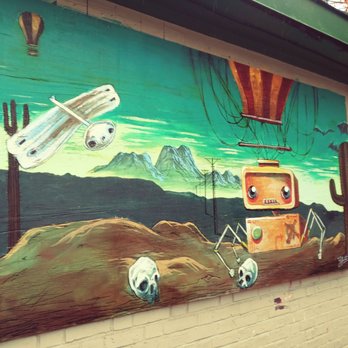 Artists Row - A mural painted on a brick wall depicts a cartoon-style robot with large eyes, skulls, cacti, and mountains in the background, with a sky and power lines. - Public Art Near Me - Salem, Massachusetts