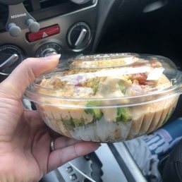 Build Your Own Poke Bowl - A person holds a plastic poke bowl with a clear lid inside a vehicle, showing salad ingredients. The car's dashboard and gear shift are visible in the background.