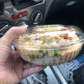 Poke Bros - Build Your Own Poke Bowl - A person holds a plastic poke bowl with a clear lid inside a vehicle, showing salad ingredients. The car's dashboard and gear shift are visible in the background. - Poke Near Me - Dover, Delaware