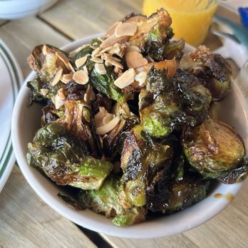 Sweet Chick - Brussel Sprouts - A white bowl contains roasted Brussels sprouts with slightly charred leaves, topped with sliced almonds. The bowl is on a wooden table, with a glass of orange juice and other dishes visible in the background, indicating a meal setting. - Southern Near Me - Brooklyn, New York