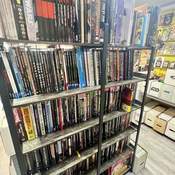 Adam's Action Figures & Comics - A metal shelving unit in a retail store holds a large collection of comic books and graphic novels, organized on multiple shelves. The titles include popular series such as Star Wars, Marvel, and other graphic novels, with some books stacked vertically and others horizontally. The store environment features additional merchandise and storage boxes visible in the background, indicating a specialty comic book shop. - Comic Books Near Me - Rootstown, Ohio