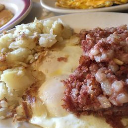 A breakfast plate includes crispy hash browns, two fried eggs with runny yolks, and a portion of corned beef hash with diced potatoes and meat. Additional dishes are visible in the background, suggesting a hearty breakfast setting.