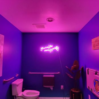 Kaliburger - The ladies - The image shows a small room with purple-painted walls, decorated with a neon sign that says 'you're really pretty' in pink. There is artwork on the left wall, a toilet in the corner, and a potted plant on the right side. A wooden sign on the right wall reads 'HELLO BEAUTIFUL.' The room has a ceiling vent and a small wall-mounted heater or panel. - Burgers Near Me - St. Augustine, Florida