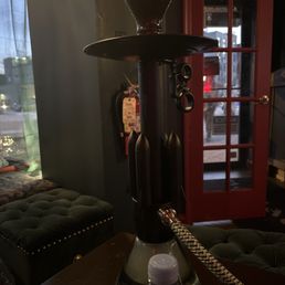 Love 66 - A black hookah with a glass base and a patterned hose is set on a table inside a lounge, with a red door and seating visible in the background.