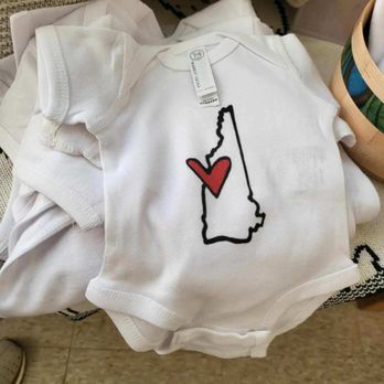 Soul Source - Eco-sustainable local baby products - A white baby onesie with short sleeves displays a black outline of the state of Maine, with a red heart positioned within the outline. The onesie is laid flat on a surface, surrounded by other clothing or fabric items, and has a tag attached near the neckline. The image emphasizes the design and the soft fabric of the garment. - Lighting Stores Near Me - Bradford, New Hampshire