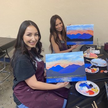Paint & Sip Studio Temecula - Two women participate in a paint and sip class, each holding a landscape painting of mountains and a sunset, with painting supplies on the table. - Paint & Sip Near Me - Temecula, California