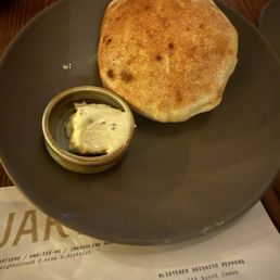 A round, dark-colored plate contains a large, golden-brown naan bread and a small dish of butter or ghee. The naan appears freshly baked with some darker spots, indicating charring. The dish of butter is partially melted and placed on a piece of paper or menu underneath the plate. The background shows a wooden table surface and part of a menu with food descriptions.