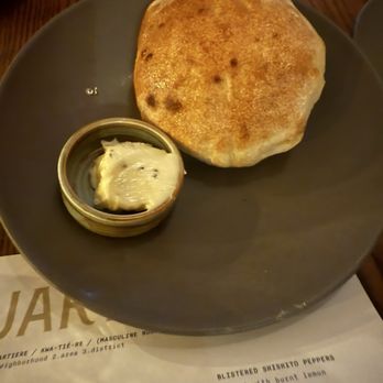 Quartiere - A round, dark-colored plate contains a large, golden-brown naan bread and a small dish of butter or ghee. The naan appears freshly baked with some darker spots, indicating charring. The dish of butter is partially melted and placed on a piece of paper or menu underneath the plate. The background shows a wooden table surface and part of a menu with food descriptions. - Pizza Near Me - Stamford, Connecticut