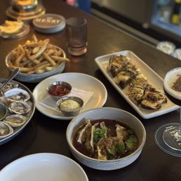 Short Rib Potstickers - A dining table features oysters on a tray, French fries, sushi rolls, and a cocktail, with various condiments and glasses.