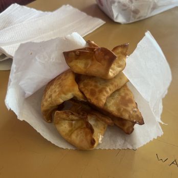 101 Drive-In - The image shows a small, white paper container filled with fried wontons that have a golden-brown, crispy exterior. The wontons are arranged in a slightly overlapping manner, with some sauce drizzled on top. The container is placed on a light-colored table, with a few napkins and a partially visible paper bag in the background. The setting appears to be casual, possibly at a restaurant or food stand. - Burgers Near Me - Willits, California