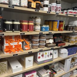 A gourmet purveyors dream - Shelves filled with various products including jars of jam, gift boxes with ribbons, and decorative items such as butterfly and floral designs. The products are arranged in an organized manner, with price tags visible. The store appears to sell gourmet food items, gifts, and decorative products, with multiple shelves holding different categories of items.